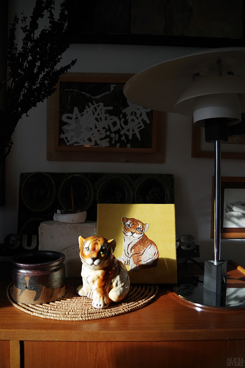 En porslinsfigur i form av en tigerunge framför ett broderat porträtt med samma motiv. Porträttet är handsytt med bomullstråd på gult linnetyg. Ur projektet Broderade porslinsdjur av Alicia Sivertsson.