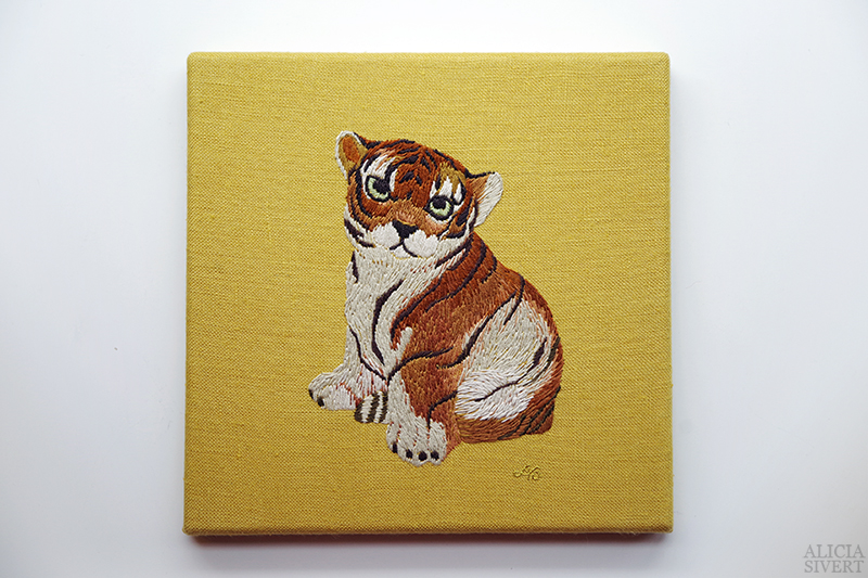 Ett broderat porträtt med motiv av en söt tigerunge som tittar under lugg. Porträttet är handsytt med bomullstråd på gult linnetyg och signerat i guldtråd. Ur projektet Broderade porslinsdjur av Alicia Sivertsson.